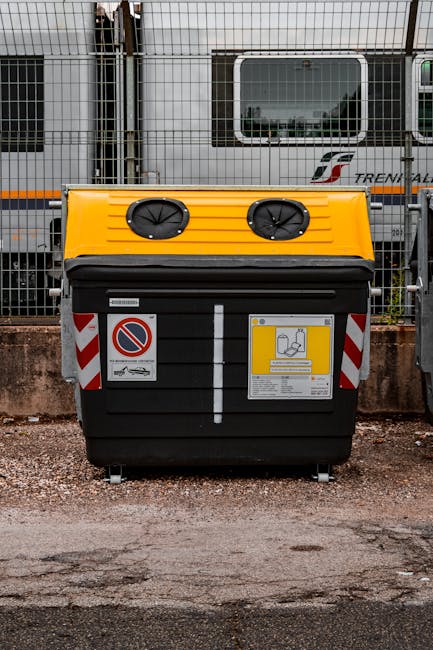 At a modern train station platform, a worker dressed in a high-visibility vest, dark clothing, and a beanie is standing beside a wheeled trolley loaded with various bags and loose waste materials, inc