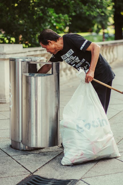 A black wheelie bin labeled 'St. John's' is positioned on the edge of a pavement beside a quiet, dimly lit street at night. The bin's lid is open, revealing a disorganized collection of mixed waste, i